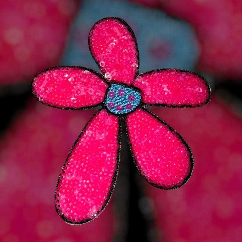 Puede incluir: Primer plano de una flor decorativa con cinco p&eacute;talos de color rosa brillante, cada uno cubierto de lentejuelas. El centro es azul con un patr&oacute;n de puntos y los bordes est&aacute;n delineados en negro.