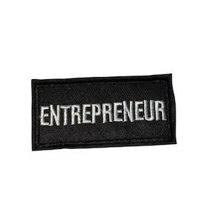 May include: A black rectangular patch featuring the word "ENTREPRENEUR" embroidered in white. The patch is designed with a simple, clear aesthetic, ideal for attaching to garments or accessories. The text is centrally positioned.