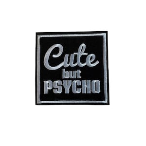 May include: A black square patch with the words "Cute but PSYCHO" embroidered in silver thread. The patch has a silver border, designed to be sewn or ironed onto clothing or accessories.