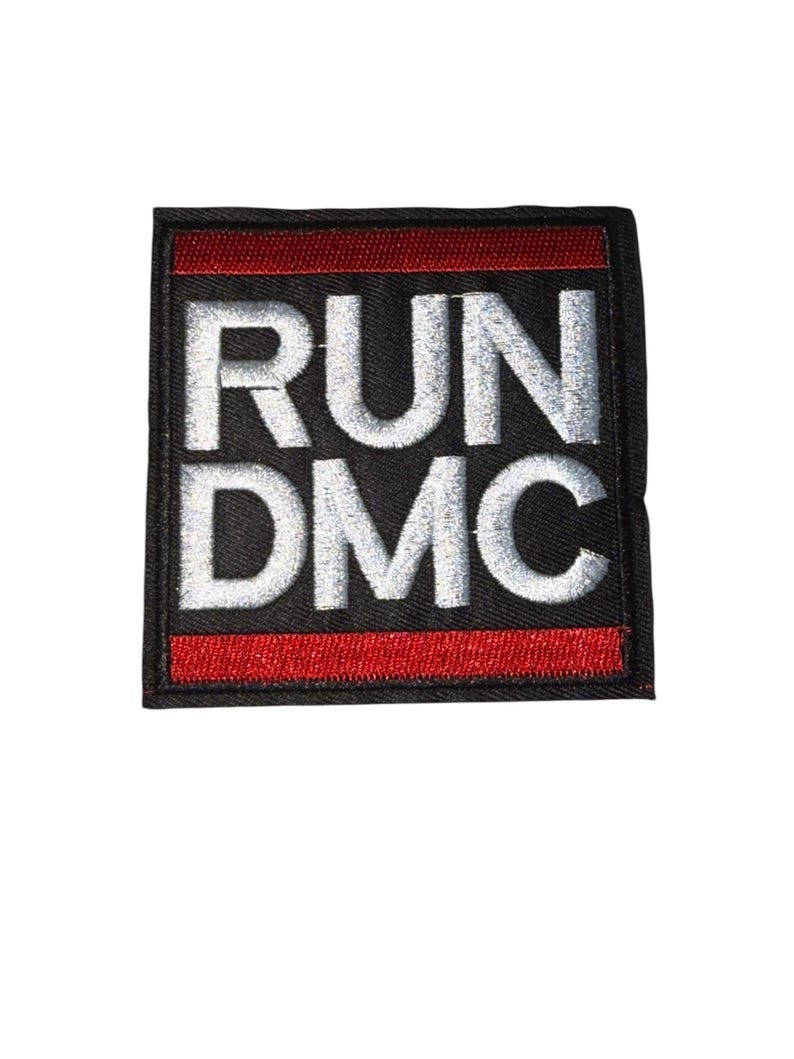 May include: Embroidered patch featuring the text "RUN DMC" in white, bold, capital letters. The text is centered on a black square with a red border. The patch is likely for clothing or accessories.