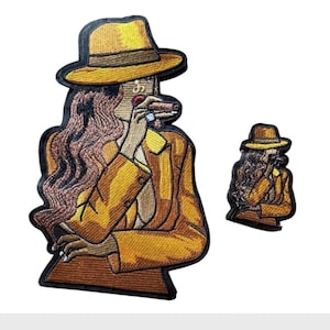 May include: Two embroidered patches featuring a figure wearing a yellow fedora and a matching yellow blazer. The figure has long wavy brown hair, is holding a brown cigar to their lips, and is outlined in a thick black border.