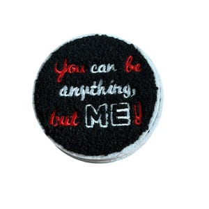 May include: A circular black embroidered patch featuring the red and white stitched text "You can be anything, but ME!". The design has a textured, fuzzy surface with a white border edge, showcasing detailed needlework on a round fabric base.