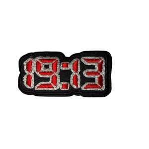 May include: Embroidered patch displaying the numbers "19:03" in a digital clock font. The red numbers have white stitching and a black outline. The patch has a black border, suitable for application on clothing or accessories.