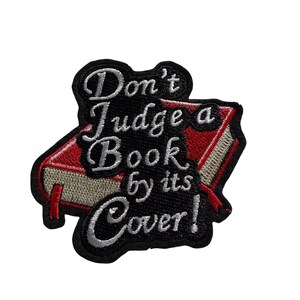 Könnte beinhalten: Gestickter Aufnäher mit einem roten und beigen Buch und dem Text "Don't Judge a Book by its Cover!" in weißer Schrift auf schwarzem Hintergrund. Zum Aufnähen oder Aufbügeln auf Stoff.