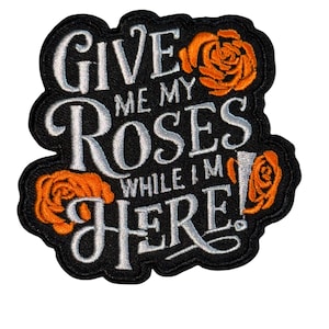 Pode incluir: Patch bordado com o texto "GIVE ME MY ROSES WHILE I'M HERE!" em escrita branca sobre fundo preto. Ilustrações de rosas laranja são colocadas ao redor do texto. O patch tem uma borda preta.