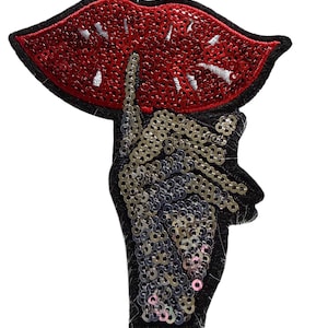 May include: A sequined patch featuring a red lip design with a black outline. A hand with a finger to the lips is also part of the design. The hand and lips are embellished with sequins in silver and red.
