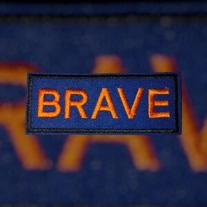 K&ouml;nnte beinhalten: Ein rechteckiger Aufn&auml;her mit dem Wort "BRAVE" in orangefarbenem Faden auf blauem Hintergrund gestickt. Der Aufn&auml;her ist mit einem schwarzen Rand umrandet. Der Hintergrund hat ein verblasstes orangefarbenes Design.