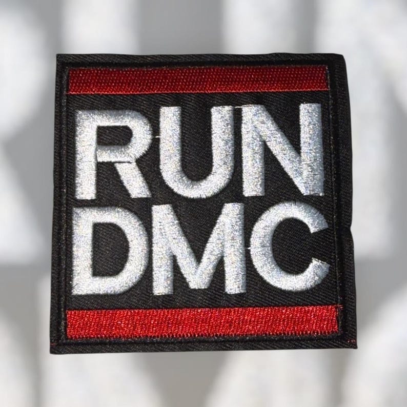 May include: A square black patch with the words "RUN DMC" embroidered in silver thread. The patch has a red stripe at the top and bottom. The patch is likely for clothing or accessories.