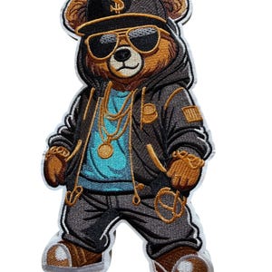 May include: Embroidered patch featuring a brown bear wearing a black hoodie, sunglasses, and a cap with a dollar sign. The bear has gold jewelry and is dressed in a hip-hop style. The patch has a white border.