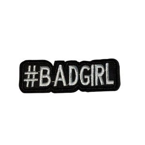 Peut inclure: Un écusson brodé noir avec le texte blanc "#BADGIRL". L'écusson est de forme rectangulaire avec des coins arrondis. Le texte est en gras, en police sans-serif.