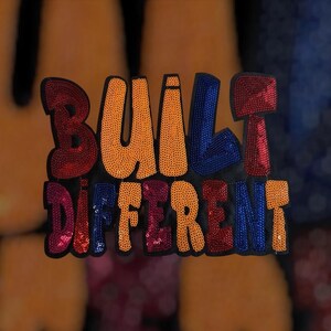 以下が含まれることがあります： カラフルなスパンコールで刺繍された「BUILT DIFFERENT」の文字が入った黒いパッチ。文字はオレンジ、赤、青、ピンク。衣類やアクセサリー用のパッチです。