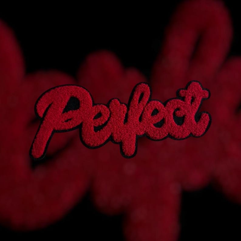May include: Red embroidered patch with the word "Perfect" in a cursive font. The patch has a black border and a textured, fuzzy appearance. The background is a blurred red color, with a black backdrop.