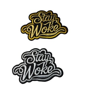 May include: Two embroidered patches with the phrase "Stay Woke". One patch features gold lettering and trim on a black background. The other patch has white lettering and trim on a black background. Both patches have a cloud-like shape.