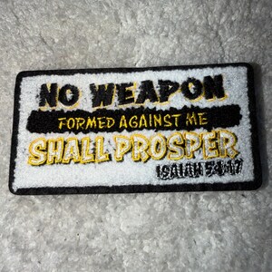 May include: Embroidered patch with the text "NO WEAPON FORMED AGAINST ME SHALL PROSPER" in black and yellow lettering. The patch has a white background with a black border and the scripture reference Isaiah 54:17.