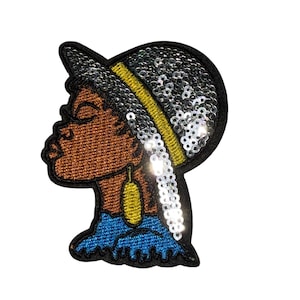 Peut inclure: Écusson brodé représentant le profil d'une personne à la peau brune, portant un chapeau noir orné de paillettes argentées et d'une bande jaune. La personne porte une boucle d'oreille dorée et un haut bleu. L'écusson est bordé de noir.