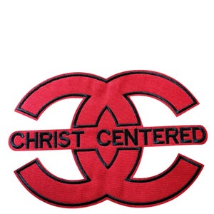 Puede incluir: Un parche bordado de color rojo con un dise&ntilde;o circular entrelazado estilizado y contorno negro. El texto CHRIST CENTERED est&aacute; bordado en letras may&uacute;sculas negras en el centro del dise&ntilde;o, dividiendo horizontalmente las dos formas entrelazadas.