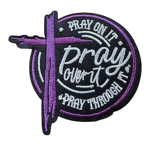 May include: A circular black embroidered patch featuring a purple cross and white text that reads "PRAY ON IT," "pray over it," and "PRAY THROUGH IT." The design includes a purple border and a textured finish with detailed stitching throughout the piece.