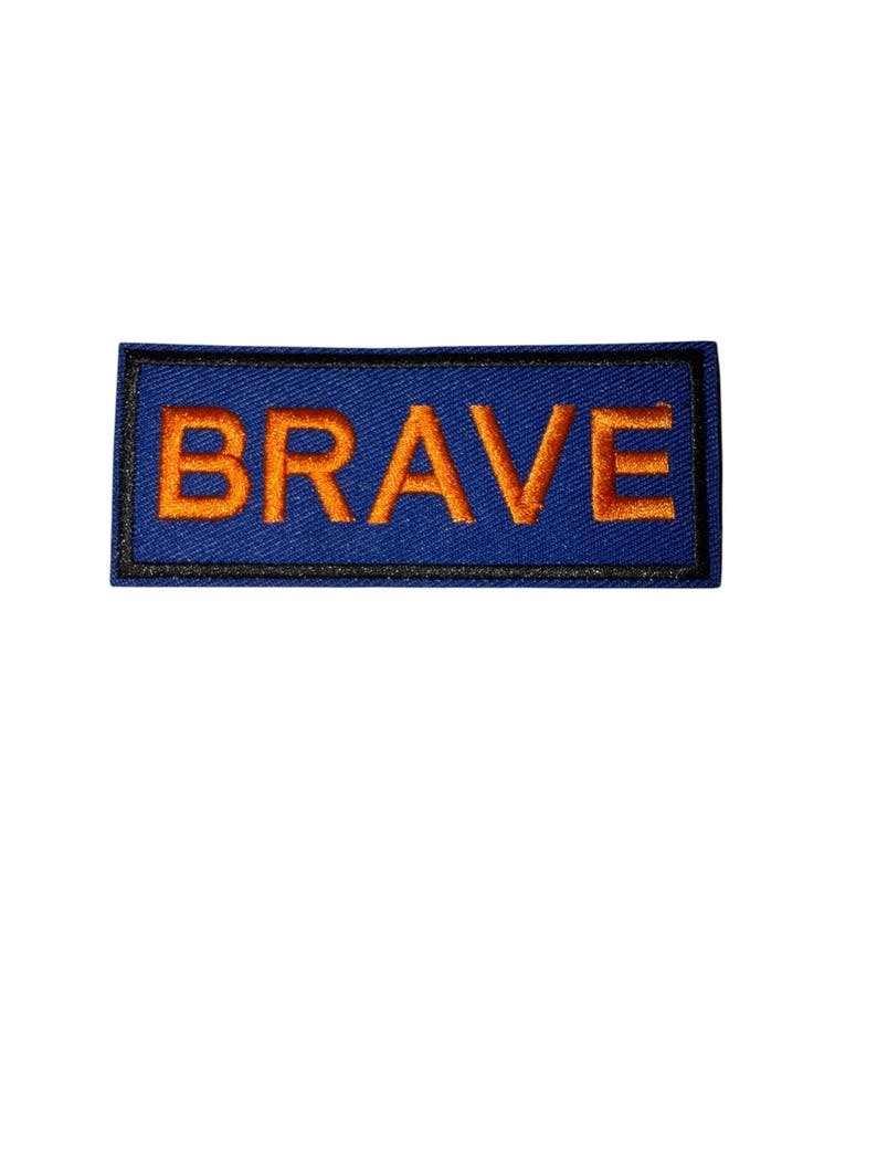 K&ouml;nnte beinhalten: Ein rechteckiger Aufn&auml;her mit dem Wort "BRAVE" in orangefarbener Stickerei auf blauem Grund. Der Aufn&auml;her hat einen schwarzen Rand und ist zum Aufn&auml;hen auf Kleidung oder Accessoires gedacht.
