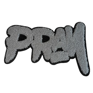 Può includere: Una toppa grigia, strutturata, con la scritta "DREW" in un carattere stilizzato. Le lettere sono bordate di nero, creando un forte contrasto sullo sfondo grigio. La toppa è probabilmente destinata a vestiti o accessori.