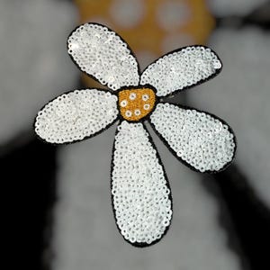 Puede incluir: Una flor de margarita blanca con lentejuelas, centro amarillo y contorno negro. La flor tiene cinco p&eacute;talos y probablemente sea una aplicaci&oacute;n o adorno para ropa o manualidades.