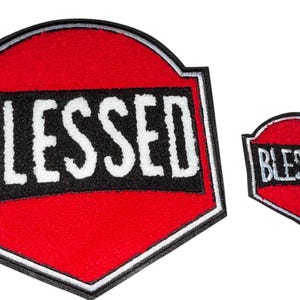 Peut inclure: Deux écussons brodés avec le mot "BLESSED". Le plus grand est rouge avec un bord noir et blanc, et le mot "BLESSED" en lettres blanches sur un rectangle noir. Un plus petit écusson assorti est également présent.