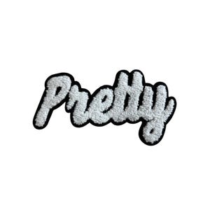 May include: A white, fluffy patch with the word "Pretty" in a cursive font. The text is outlined in black, creating a bold contrast against the white background. The patch has a textured, fuzzy appearance.