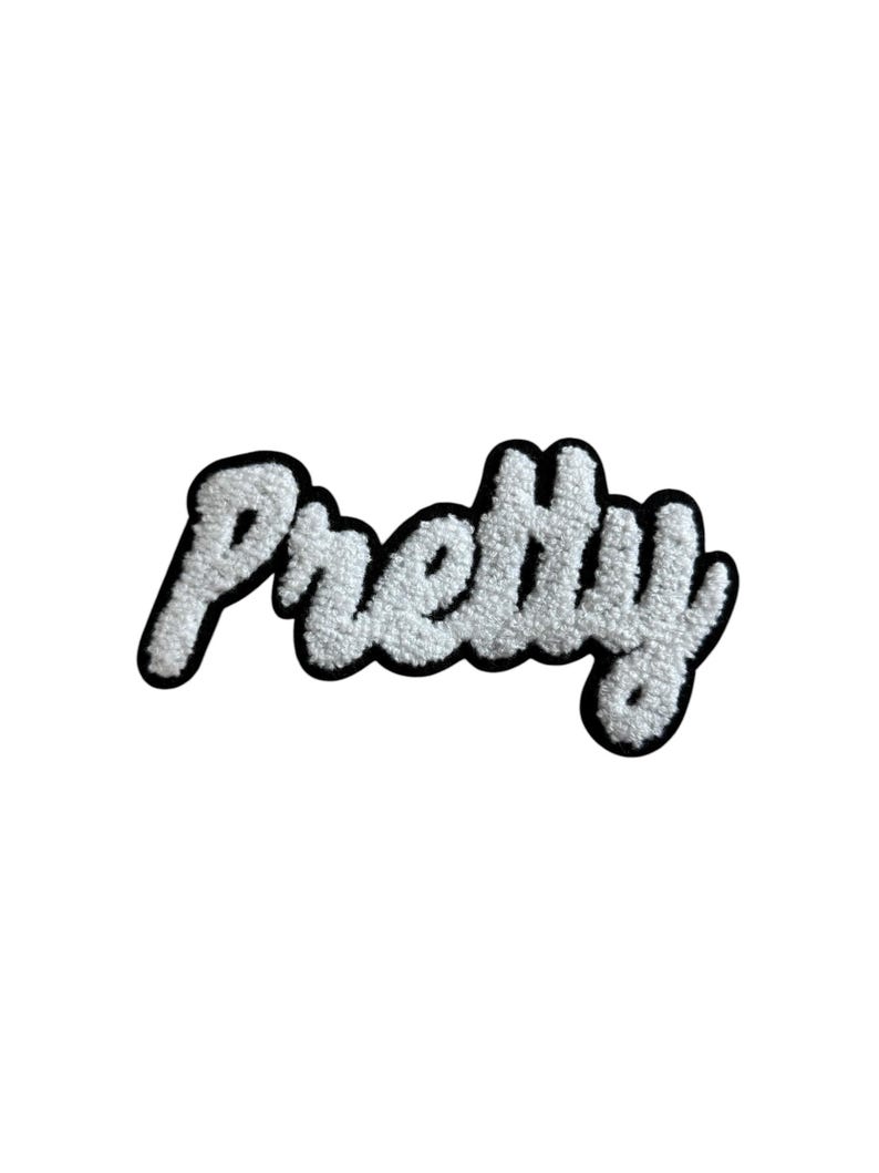 May include: A white, fluffy patch with the word "Pretty" in a cursive font. The text is outlined in black, creating a bold contrast against the white background. The patch has a textured, fuzzy appearance.