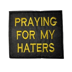 Könnte beinhalten: Schwarzer, bestickter Aufnäher mit dem gelben Text "PRAYING FOR MY HATERS". Der Aufnäher hat eine rechteckige Form mit einem schwarzen Rand.