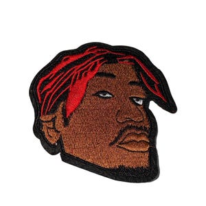 May include: Embroidered patch featuring a stylized portrait of a person with brown skin, red hair, and a black outline. The patch has a detailed design with visible stitching, suitable for application on clothing or accessories.