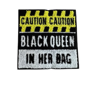 Op de afbeelding: Een zwart-wit patch met de tekst "CAUTION CAUTION" in geel boven "BLACK QUEEN IN HER BAG" in wit. De patch heeft een getextureerde, geborduurde uitstraling.