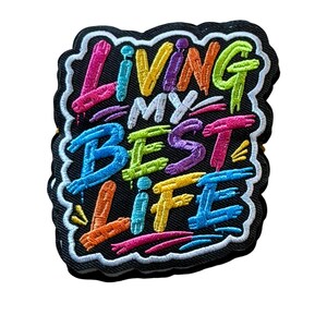 May include: A black embroidered patch with a white border featuring the multi-colored graffiti-style text "LIVING MY BEST LIFE" in pink, yellow, purple, green, and blue threads. The design has a textured appearance with visible stitching.