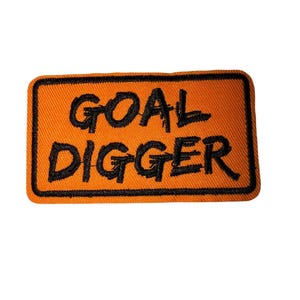 Puede incluir: Parche rectangular naranja con texto bordado en negro que dice "GOAL DIGGER". El parche tiene un borde negro y está sobre un fondo blanco. Adecuado para chaquetas, bolsos u otros artículos de tela.