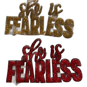 Puede incluir: Dos letreros decorativos con purpurina con la frase "She is Fearless". El letrero superior es dorado, el inferior es rojo. Ambos letreros tienen un respaldo blanco y están sobre un fondo blanco.