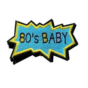 May include: An embroidered patch shaped like a jagged comic book speech bubble with a bright blue center, a yellow border, and a black outline. The text 80'S BABY is stitched in black across the center of the blue field in a bold, sans-serif font.