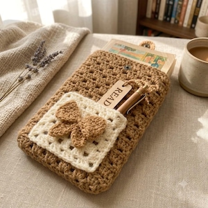 May include: A tan crochet book sleeve featuring a cream-colored square pocket adorned with a matching tan crochet bow. A wooden tag with the word READ is tucked into the pocket alongside a pen. The sleeve has a button closure at the top.