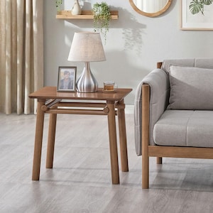 May include: A wooden side table with four tapered legs and a lower shelf, featuring a silver-toned table lamp with a beige fabric shade, a small framed photograph, and a clear glass mug containing a light brown beverage on its surface.