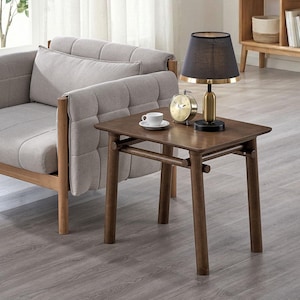 May include: A rectangular wooden side table with four cylindrical legs and horizontal support bars. On the table sits a black and gold table lamp, a gold-toned analog clock, and a white ceramic cup and saucer set on a small white plate.