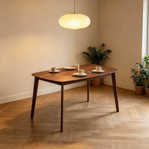 May include: A wooden dining table with a warm brown finish, set with four place settings. The table is supported by four angled legs. Above, a round, cream-colored pendant light hangs. Potted plants are in the background.