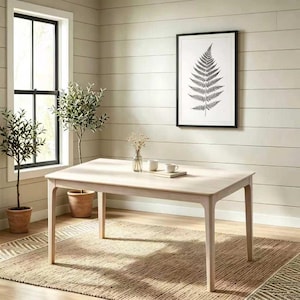 May include: A light-toned wooden dining table with four tapered legs, positioned on a woven rug. On the table surface sit two white ceramic mugs and a small glass vase with dried flowers. A framed black and white fern print hangs on the wall.