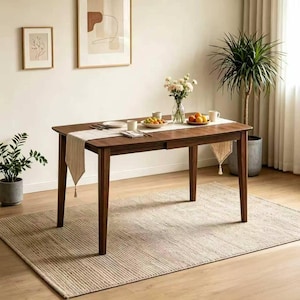 May include: A dark brown wooden dining table with a rectangular top and four legs. The table is set with plates, cups, and a runner. A beige rug is beneath the table. The room has plants and framed artwork.