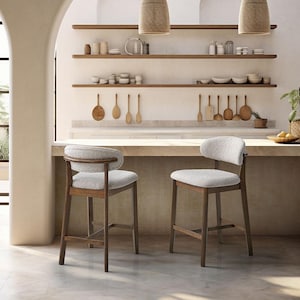 May include: Two bar stools with dark brown wooden frames and textured off-white upholstered seats and curved backrests. The stools feature four straight legs connected by horizontal support bars, positioned in front of a kitchen counter.