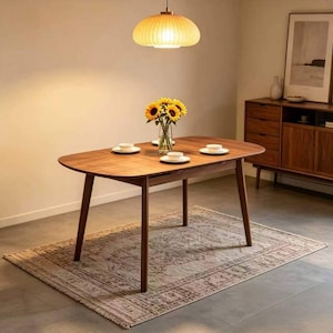 May include: A wooden oval dining table with four white teacups and a vase of sunflowers. The table is set on a patterned rug. A wooden cabinet and a hanging light fixture are also visible.