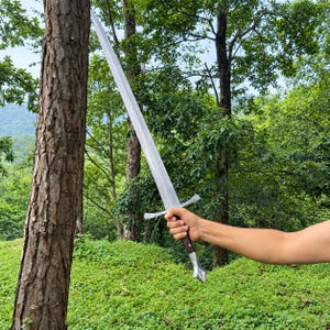May include: A long, double-edged steel sword with a pointed tip, a silver crossguard, a dark brown wooden handle, and a silver pommel. The blade features a central fuller running down the length, reflecting light against a natural outdoor setting.