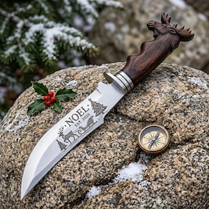 Pode incluir: Uma faca de lâmina fixa com cabo de madeira castanho-escuro esculpido em forma de cabeça de alce. A lâmina prateada apresenta uma gravação de uma rena, um trenó e árvores de Natal com o texto NOEL EST. 2026. Uma pequena bússola de latão.