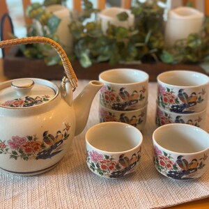 Pode incluir: Um conjunto de chá de cerâmica creme, incluindo uma chaleira com uma alça trançada e seis xícaras combinando. A chaleira e as xícaras são decoradas com motivos florais e de pássaros em vermelho, rosa e azul. O conjunto está disposto sobre uma toalha de mesa neutra.