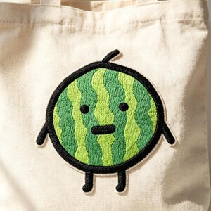 May include: A natural-colored canvas tote bag featuring a large, circular embroidered patch of a green watermelon character. The watermelon has a striped green rind, two small black eyes, a horizontal black mouth, and thin black limbs.