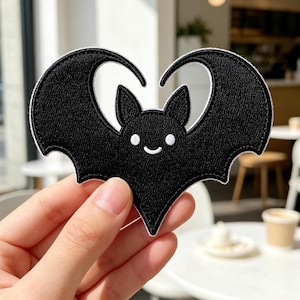 May include: A black embroidered patch in the shape of a bat with its wings curved upward to form a heart silhouette. The bat features two small white circular eyes and a simple white curved line for a mouth, all outlined with a white border.