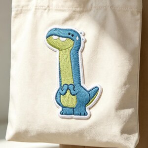 May include: A cream-colored canvas tote bag featuring a large, embroidered patch of a blue and light green cartoon dinosaur. The dinosaur has a long neck, a wide grin, and a small sweat drop on its head, centered on the front of the bag.