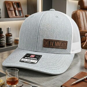 May include: A light gray trucker hat featuring a white mesh back and a rectangular brown leather patch on the front panel. The patch is embossed with the Roman numerals VI.IV.MMXIX. A black and white sticker is attached to the curved brim.