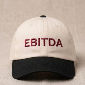 May include: A baseball cap with a cream-colored crown and a black brim. The front of the crown features the word EBITDA embroidered in dark red capital letters. The cap has a small black button at the top center and stitched eyelets.